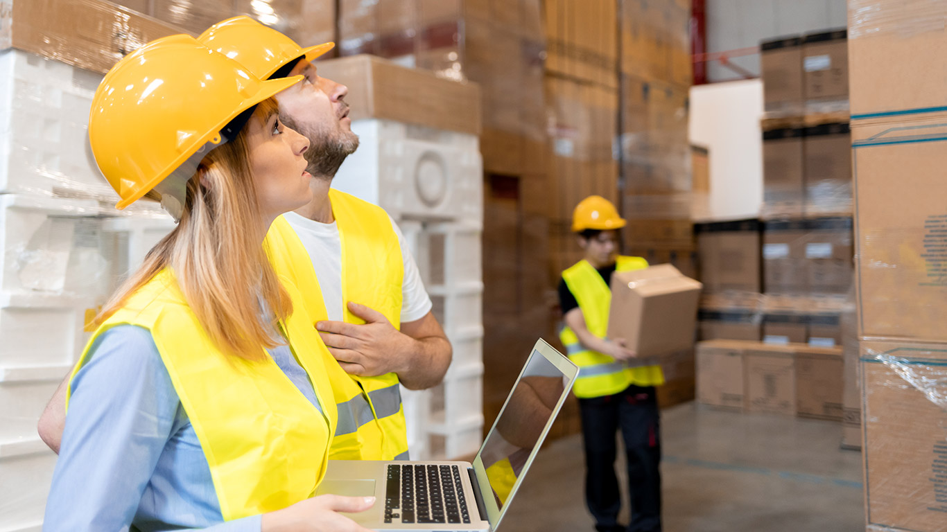 Pracownicy magazynu w żółtych kaskach spoglądający w górę.